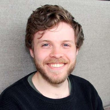 En ung mann med lysebrunt hår og skjegg smiler til kameraet mens han sitter mot en lys bakgrunn.