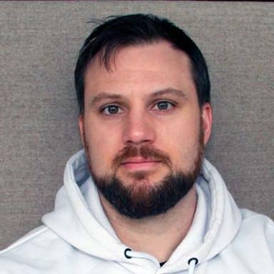 En mann med kort, mørkt hår og skjegg, iført hvit hettegenser, sitter mot en nøytral, brun bakgrunn og ser rett inn i kameraet.