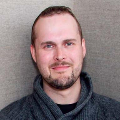 En mann med kort hår og trimmet skjegg, iført mørk genser med sjalkrage, står foran en nøytralfarget bakgrunn, ser inn i kameraet og smiler lett.