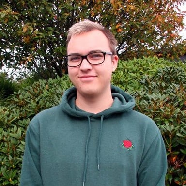 En person med briller og grønn hettegenser med et rødt roseemblem står foran grønne busker og et tre.