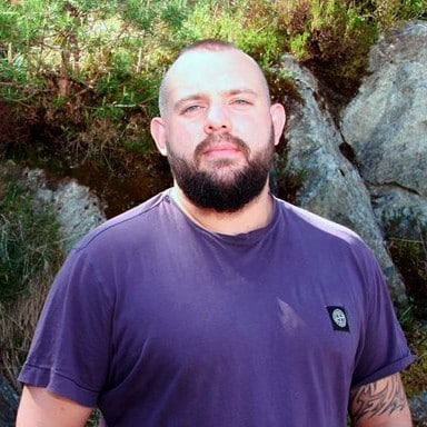 En mann med skjegg og kort hår står utendørs foran steiner og grøntområder, iført en lilla t-skjorte.