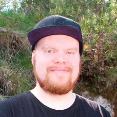 En mann med rødt skjegg og bart, iført svart lue og svart t-skjorte, står utendørs foran et grønt område.