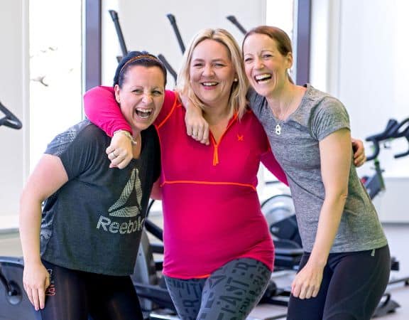 xr:d:DAFU0xRWXoo:6,j:478199360,t:22121511 Tre kvinner i sportsklær smiler og poserer sammen i et treningsstudio med treningsutstyr i bakgrunnen.
