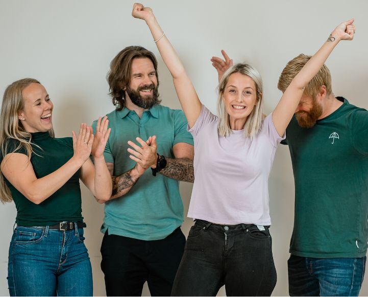 Fire voksne står sammen innendørs. Kvinnen foran løfter armene begeistret, mens de tre andre smiler og klapper, noe som tyder på feiring eller støtte.