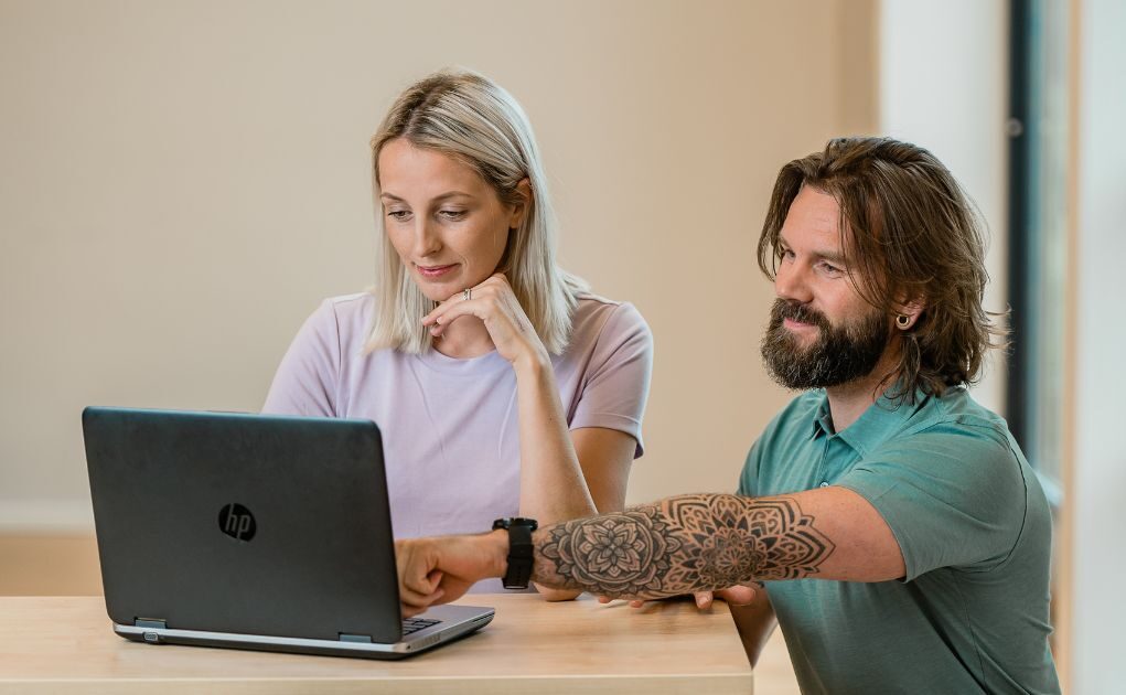 En kvinne og en mann sitter sammen ved et bord og ser på en åpen HP-laptop; mannen peker på skjermen, og begge virker fokuserte.